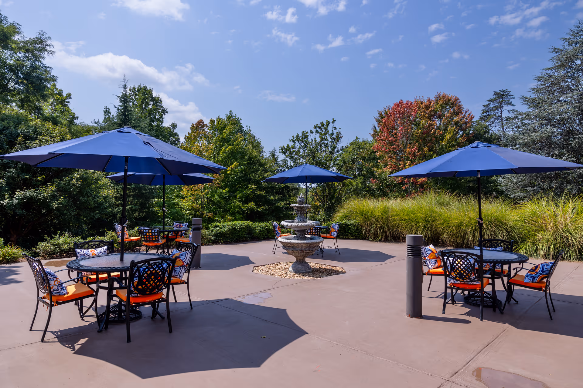 Outdoor patio area with round tables and chairs featuring orange cushions and decorative pillows. Each table is shaded by a large blue umbrella. In the center of the patio is a three-tiered stone fountain surrounded by small rocks. The patio is surrounded by green trees and bushes under a partly cloudy blue sky.