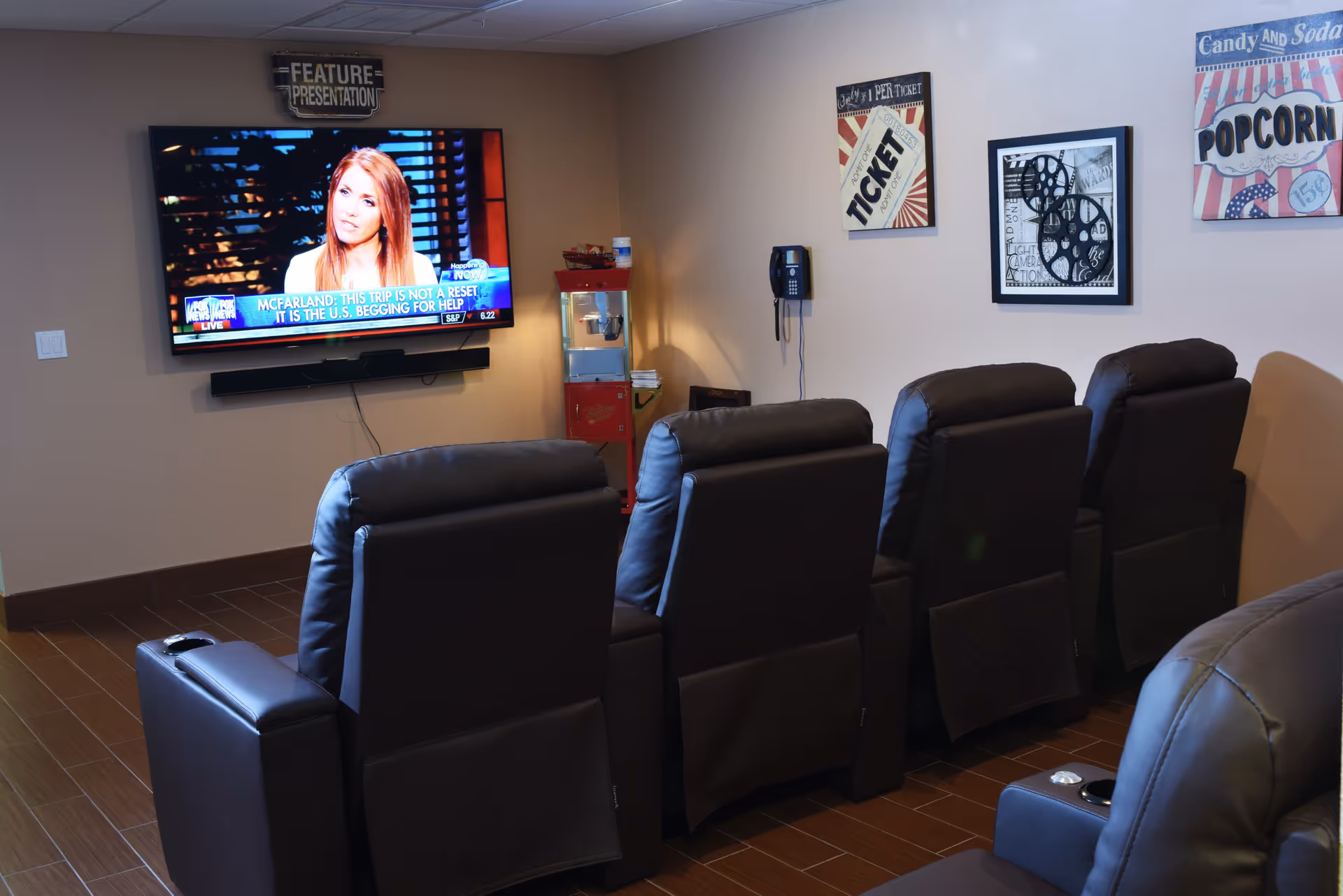 A small home theater room with five black leather recliner chairs arranged in two rows facing a wall-mounted flat-screen TV. The TV is showing a news program with a female anchor. On the wall above the TV is a sign that reads 'FEATURE PRESENTATION'. To the right of the TV is a vintage-style popcorn machine and a wall-mounted telephone. The walls have movie-themed decorations including a ticket stub, film reels, and a popcorn sign. The floor is covered with brown tiles.