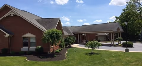 Exterior view of a brick senior living facility building with a covered entrance driveway, green lawn, small trees, and a partly cloudy sky.