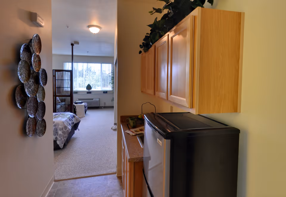 Compact kitchenette with overhead cabinets and a small refrigerator opening into a studio bedroom with a bed and large window.