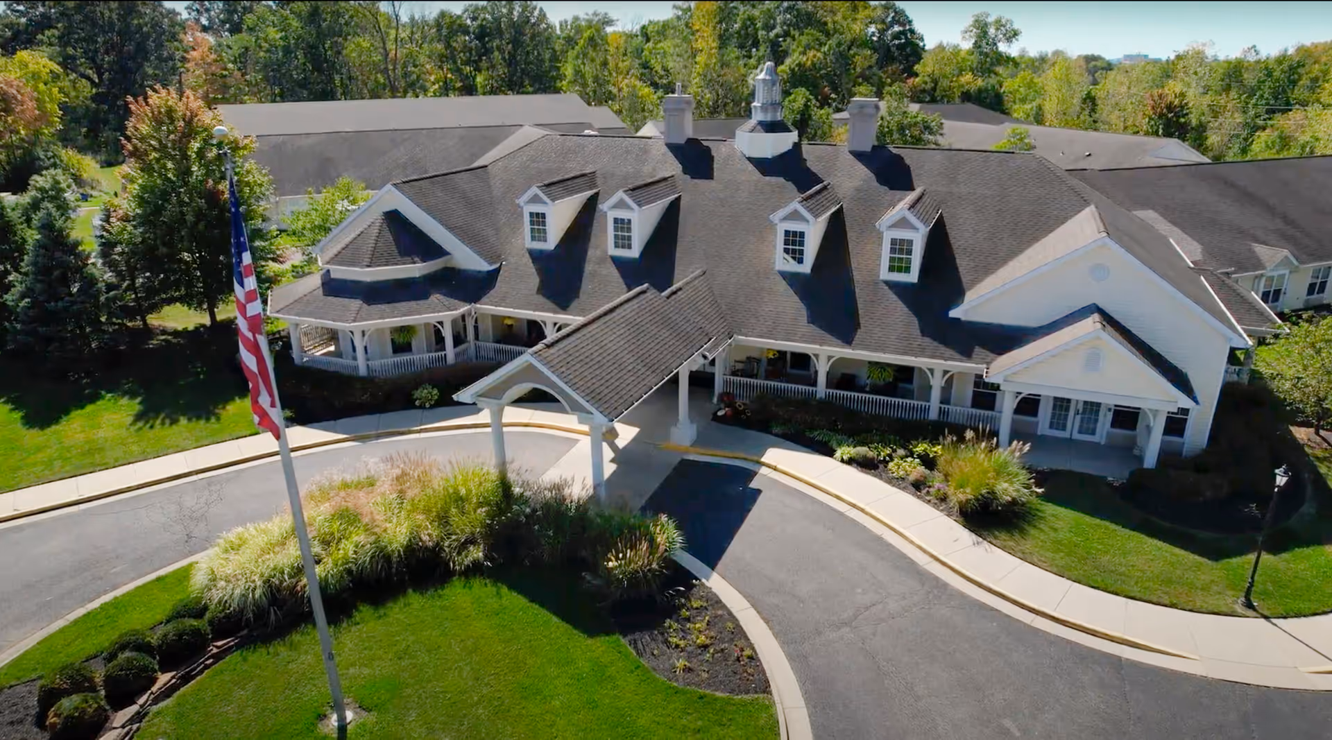 Aerial view of a large senior living facility building with a covered entrance, surrounded by well-maintained landscaping including green lawns, bushes, and trees. An American flag is visible on a flagpole near the driveway.