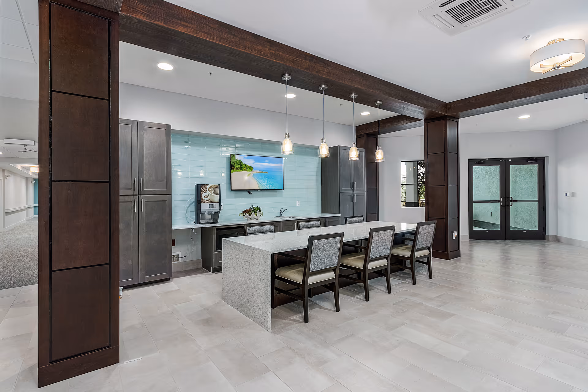 A modern communal dining area with a long rectangular table surrounded by six chairs. The room features dark wooden beams and columns, pendant lights hanging above the table, a wall-mounted TV displaying a beach scene, and a coffee machine on a countertop against a light blue tiled wall. Double glass doors and a window provide natural light to the space.