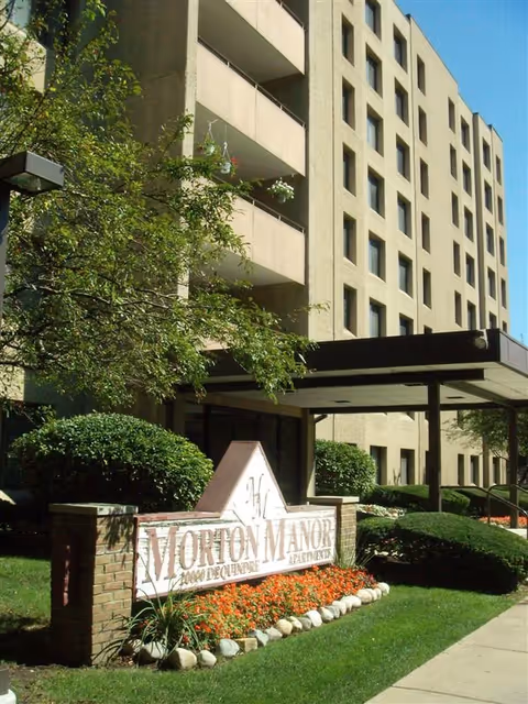 Exterior view of Morton Manor Senior Apartments building with multiple floors, balconies, and a covered entrance. The foreground shows a landscaped area with bushes, flowers, and a sign reading 'Morton Manor Apartments'.