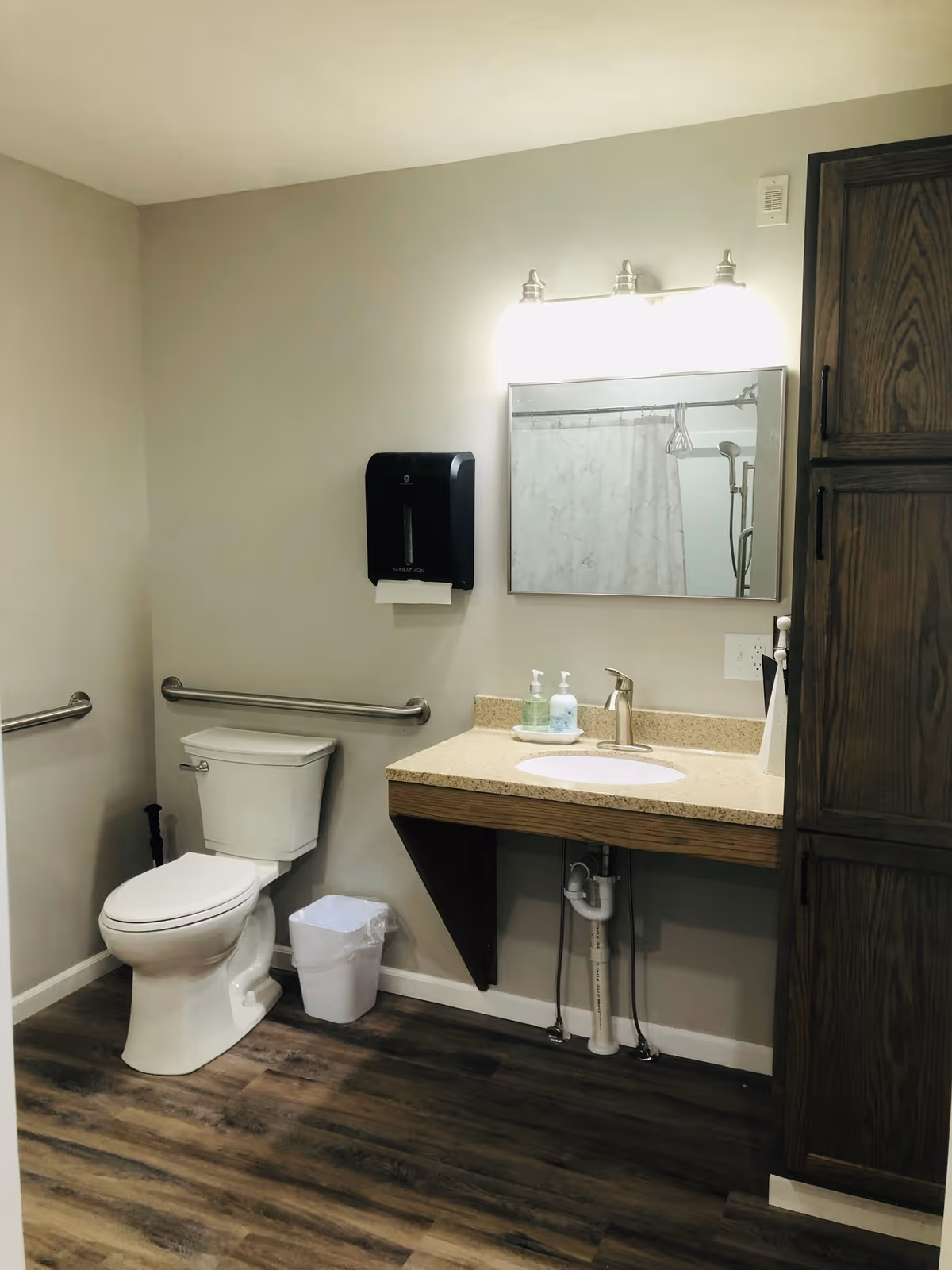 Well-lit accessible bathroom with a toilet, wall-mounted grab bars and paper dispenser, a vanity sink under a mirror, and a tall wooden cabinet.