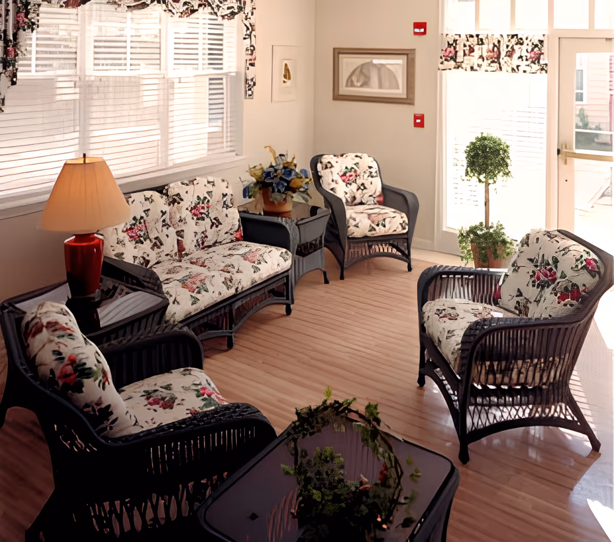 A cozy sitting area with wicker furniture including a loveseat and three armchairs, all with floral patterned cushions. There is a side table with a red lamp and a flower arrangement, a glass-top coffee table with a decorative plant, and large windows with blinds letting in natural light. A potted topiary plant is near a glass door, and framed artwork hangs on the walls.