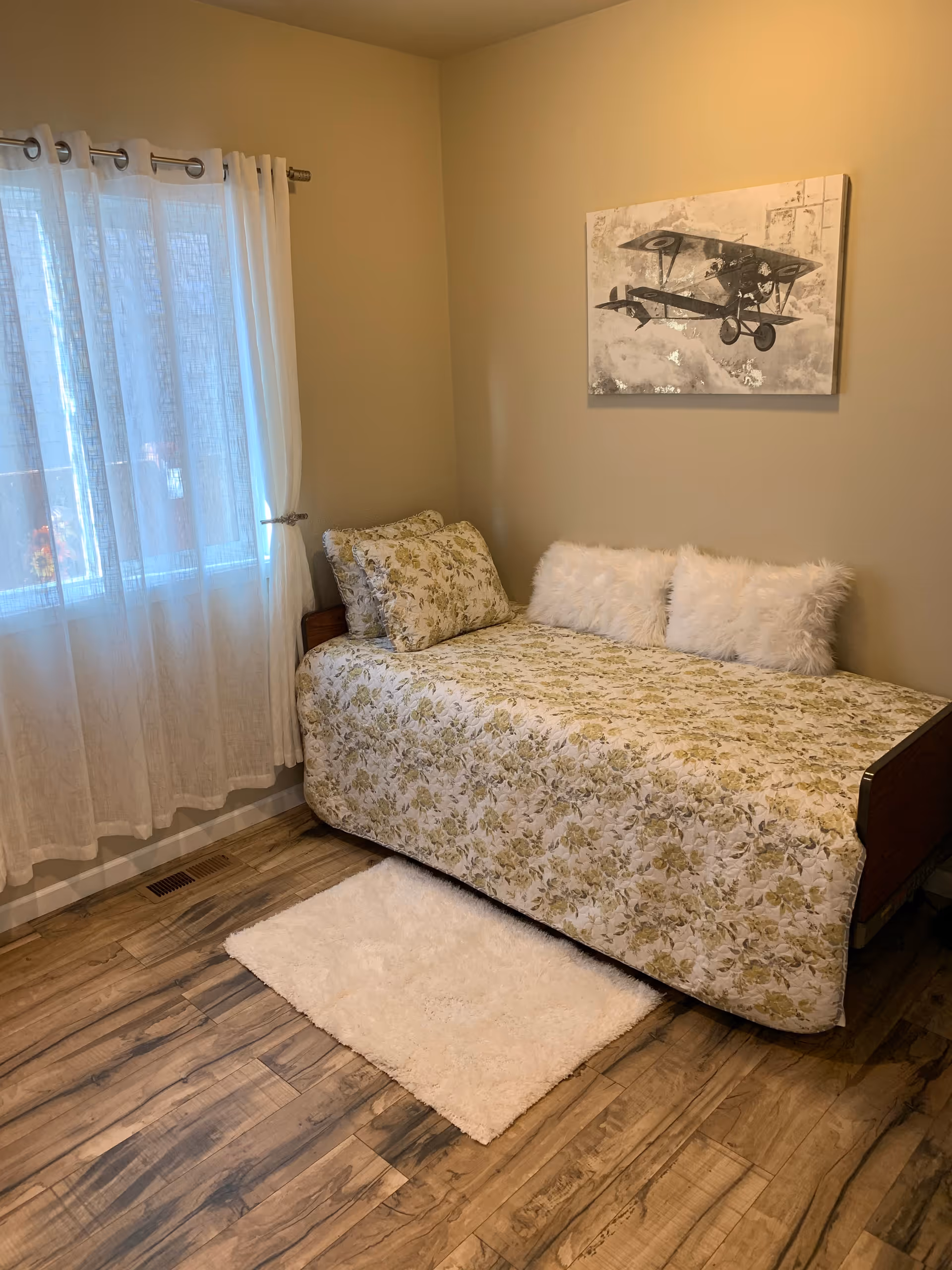 Simple bedroom with a single bed in floral bedding, fluffy pillows, a small white rug, a window with sheer curtains, and airplane wall art.