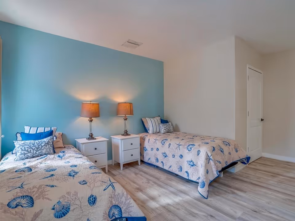 A bedroom with two twin beds featuring seashell and starfish patterned bedspreads. Each bed has multiple pillows, and there are two white nightstands between the beds, each with a lamp. The wall behind the beds is painted light blue, while the other walls are white. The floor is light-colored wood, and there is a closed white door on the right side of the room.