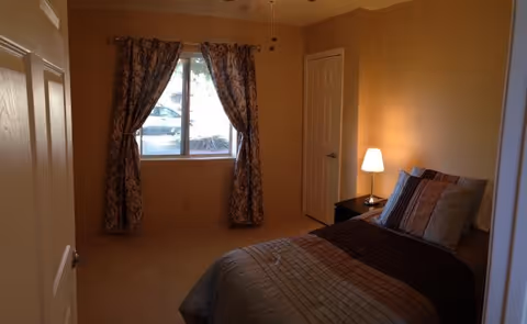 Bright bedroom with a bed, nightstand lamp, and window with patterned curtains.