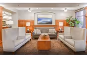A cozy living room area with two white armchairs facing each other, a gray sofa in the background, a wooden coffee table in the center, two table lamps on side tables, a large framed landscape painting on the wall, and a tall green plant near a window with curtains.