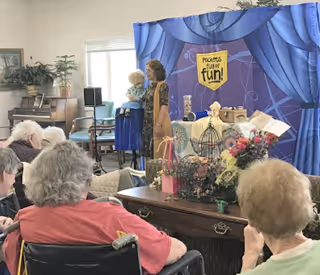 A group of elderly people seated and watching a woman holding a puppet on a stage with a blue curtain backdrop that says 'Puppets for fun!'. The setting appears to be a common area with plants, a piano, and various decorations.