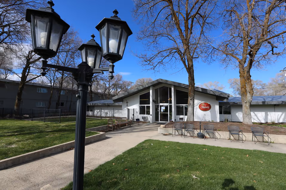 Exterior view of Aspen Assisted Living facility showing a single-story building with large windows and a peaked roof. In front of the building is a paved walkway, several chairs, benches, a black vintage-style street lamp, and leafless trees under a clear blue sky.
