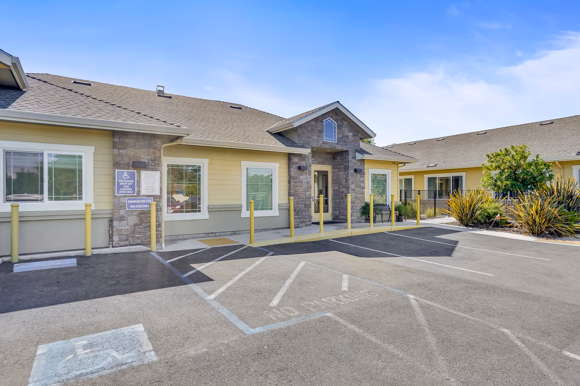 Front entrance and parking lot of a single-story senior living building with accessible parking spaces and yellow bollards.