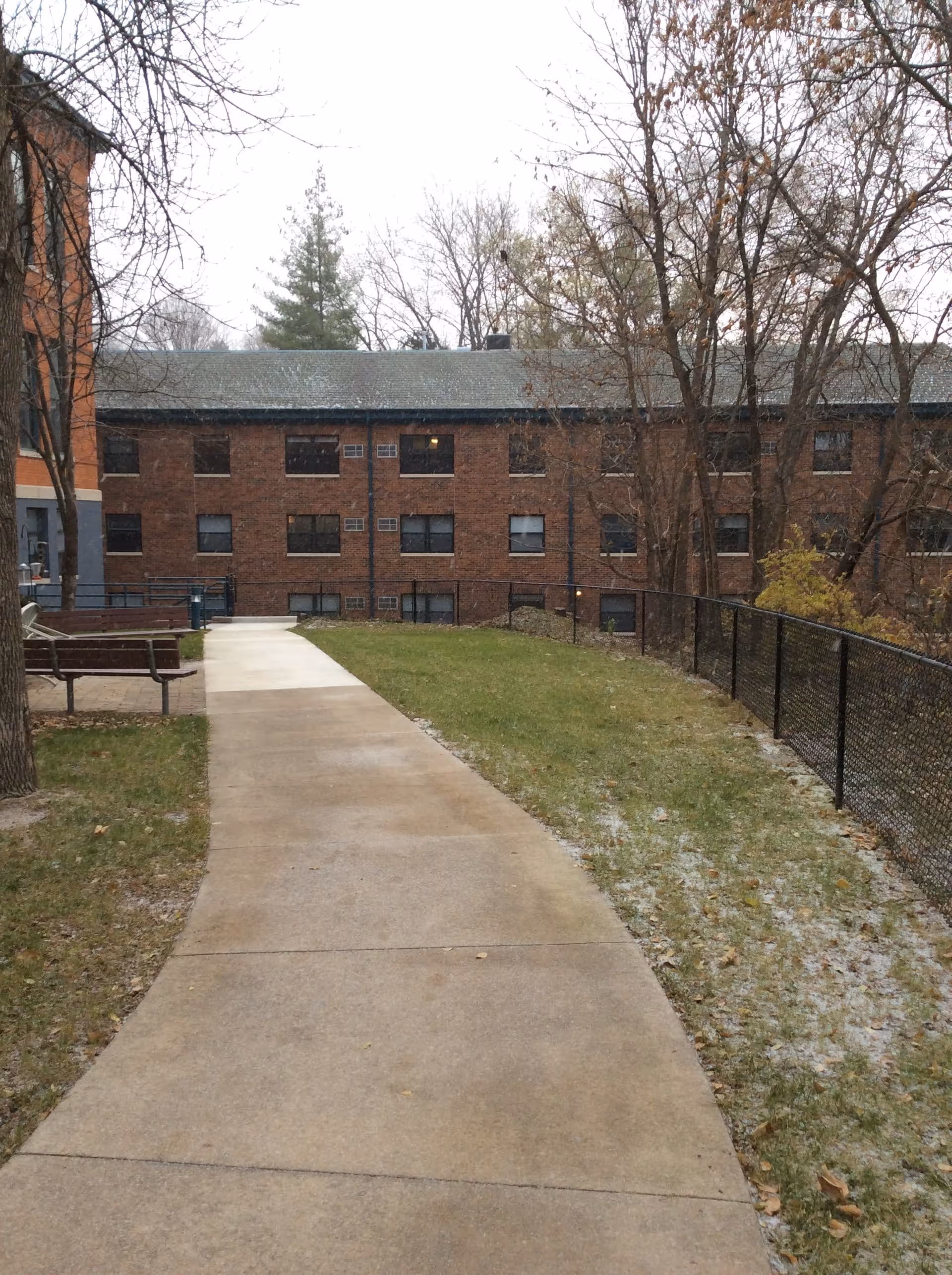 A paved walkway leading through a grassy area with a black chain-link fence on the right side and a bench on the left side. Leafless trees are visible, and a three-story brick building with multiple windows is in the background under an overcast sky.
