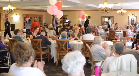 A large dining room filled with elderly people seated at round tables covered with white tablecloths. Some tables have pink and red balloons tied to chairs. The room is well-lit with chandeliers, and several people are standing or walking around, engaging with the seated guests.