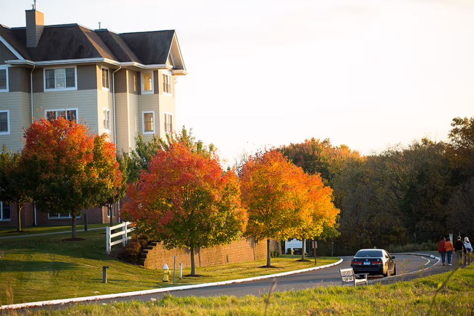 A senior living facility building with beige and brown exterior walls, surrounded by trees with vibrant autumn foliage in red, orange, and yellow. A curved road runs alongside the building with a parked car and a few people walking in the distance. The scene is set in a peaceful, natural environment during the daytime.