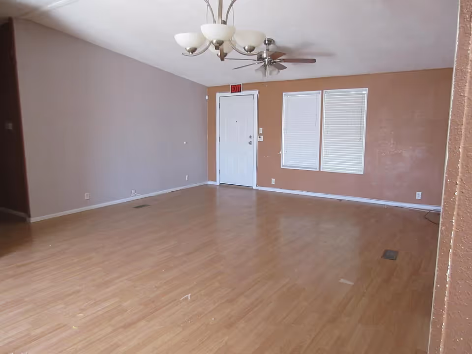 Empty room with wooden flooring, beige and brown walls, a white door with an exit sign above it, two windows with closed blinds, a ceiling fan, and a chandelier light fixture.