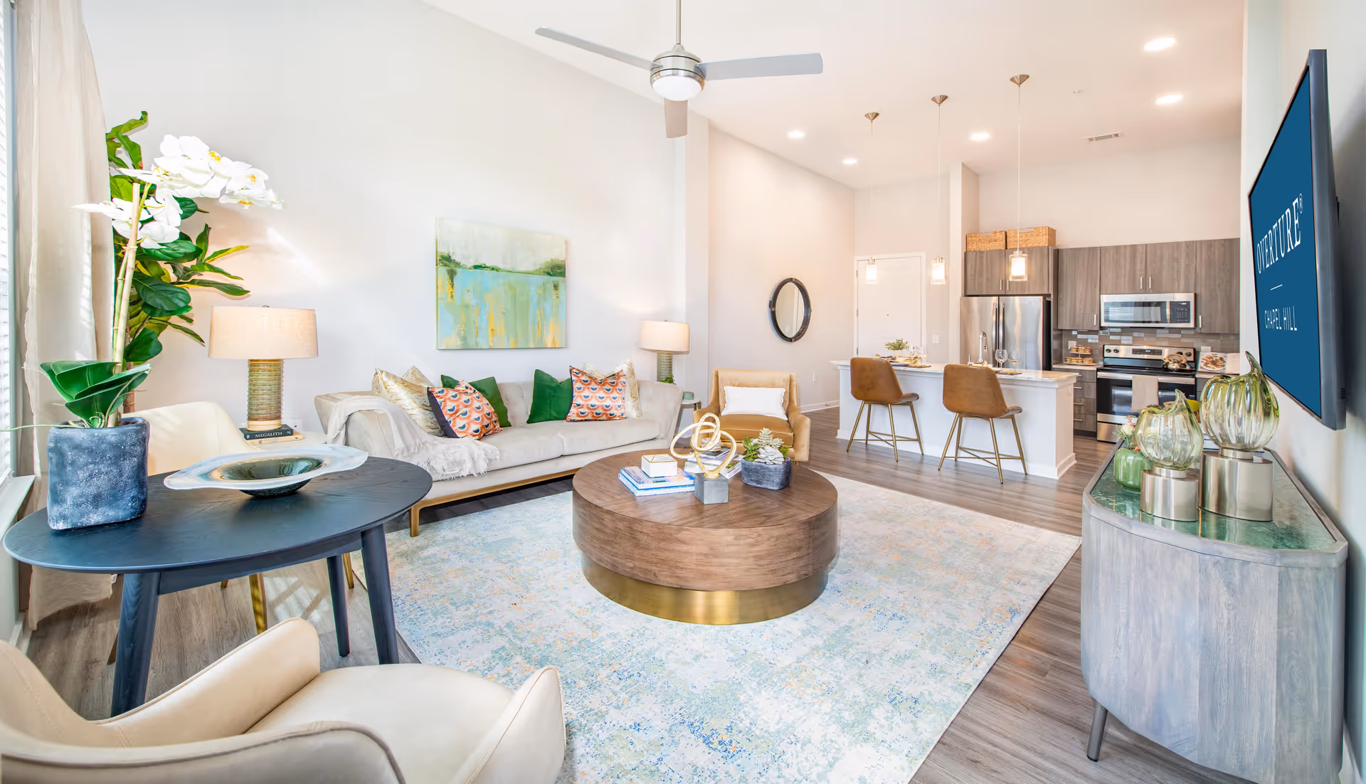 Bright open-plan living room and kitchen with a sofa, armchairs, round coffee table on a large rug, and a wall-mounted TV.