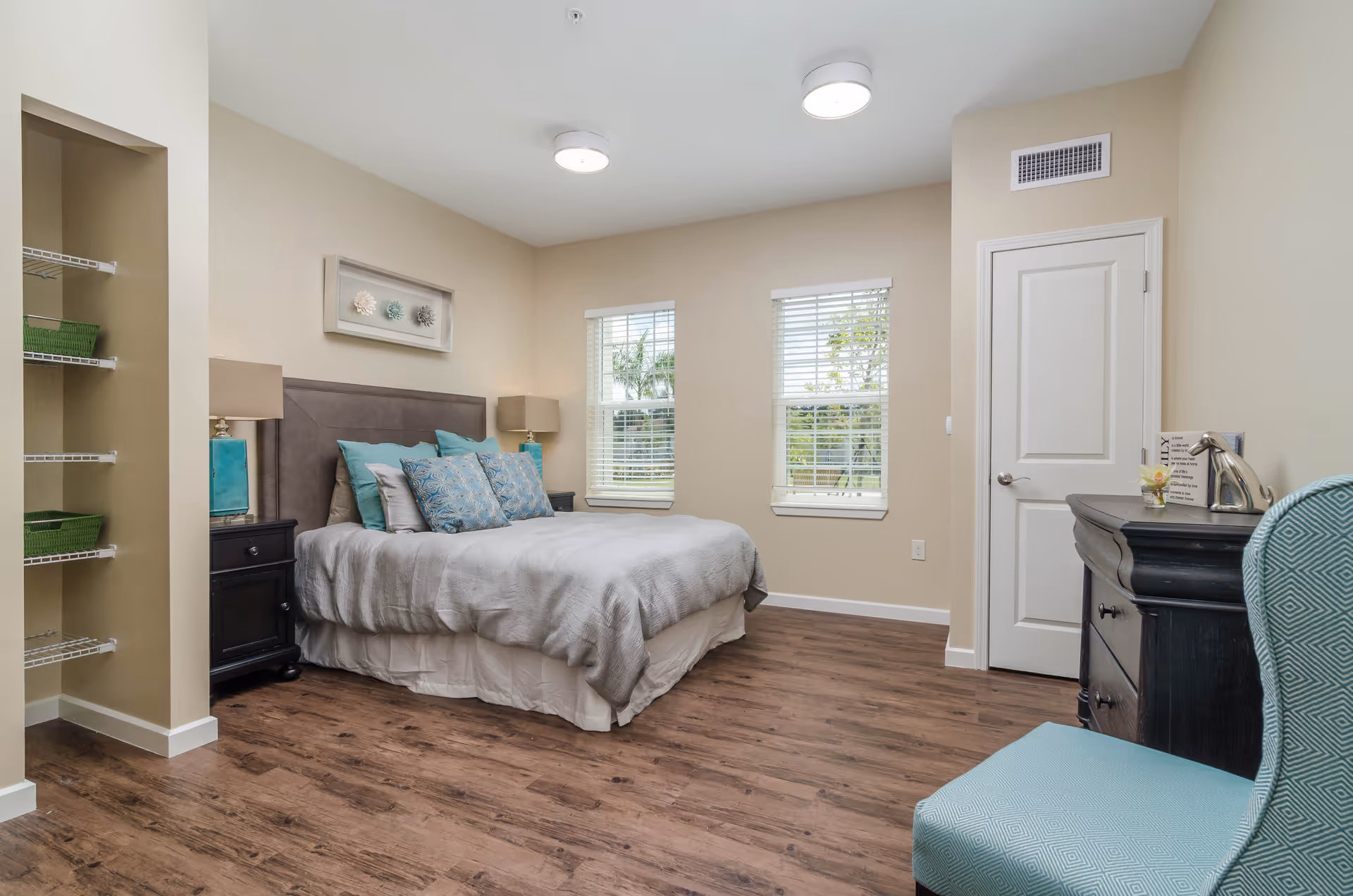 A neatly arranged bedroom with a large bed featuring multiple pillows in shades of blue and gray. There are two windows with white blinds letting in natural light, a black nightstand with a lamp on each side of the bed, a black dresser with decorative items, and a teal upholstered chair. The room has wood flooring and beige walls.