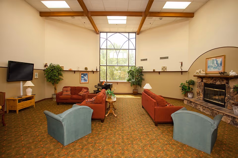 A spacious living room with high ceilings and wooden beams. The room features a large window with decorative framing, two red sofas, two blue armchairs, a stone fireplace with a mantel, a flat-screen TV mounted on the wall, a piano near the window, and several potted plants and lamps. The carpet has a patterned design, and the walls are painted in a two-tone beige color.