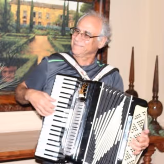 An elderly man with gray hair and glasses playing a black and white accordion indoors, with a framed painting and decorative wooden objects in the background.
