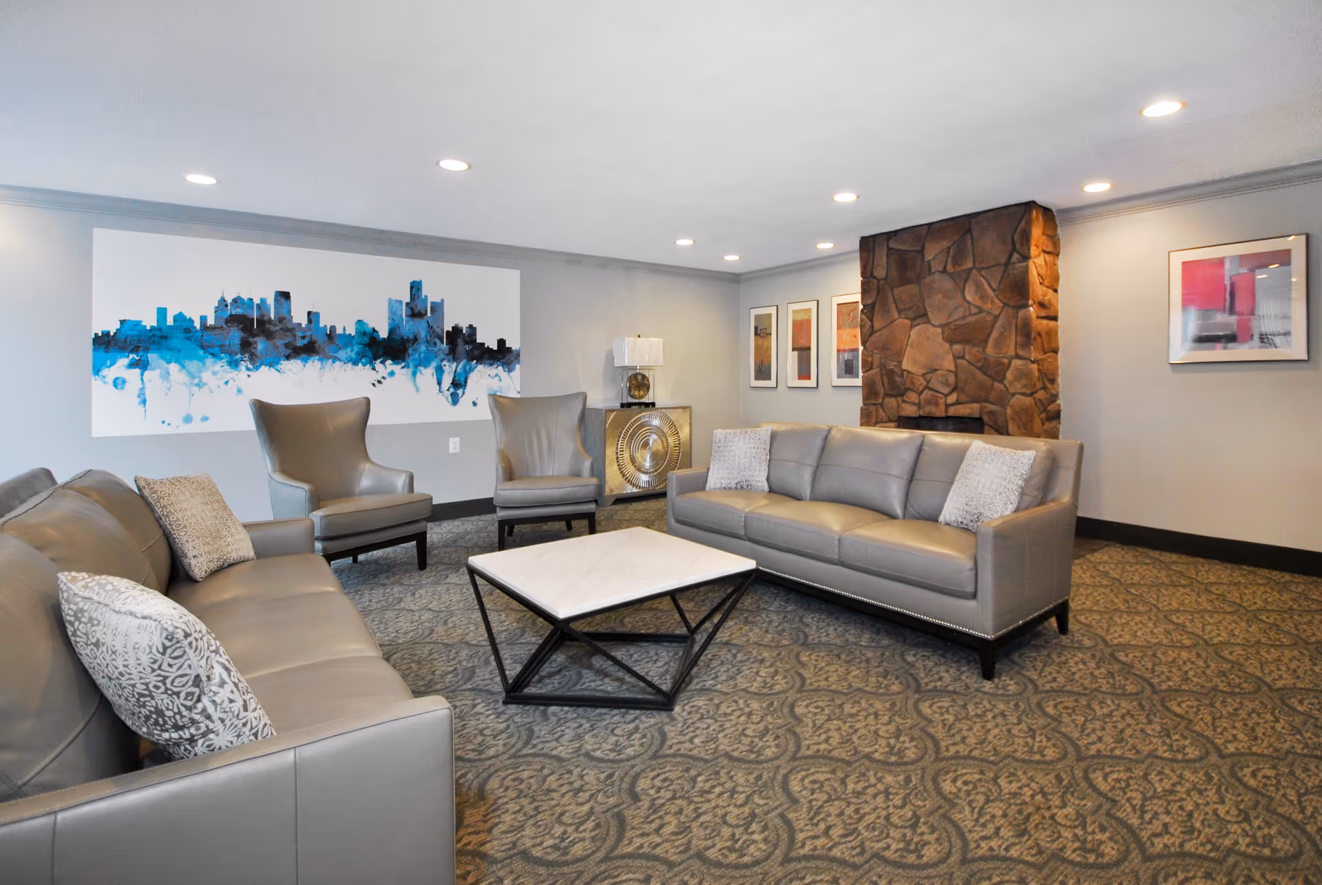 A modern living room with two gray leather sofas, two gray armchairs, a square marble coffee table with a black metal frame, a stone fireplace, and several framed artworks on the walls. The room has recessed ceiling lights and patterned carpet flooring.