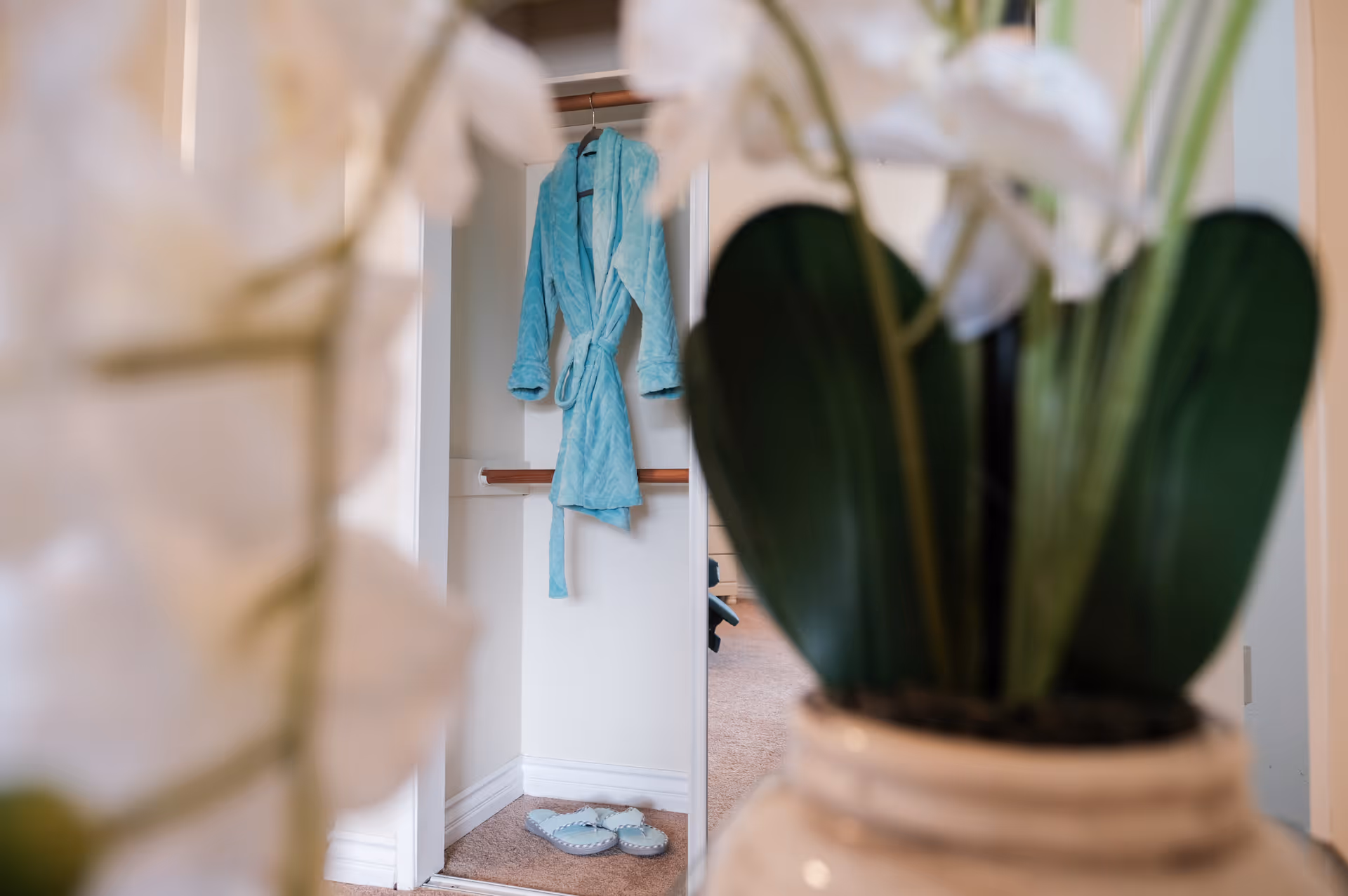 A light blue bathrobe hanging on a hook inside a small closet or alcove with a pair of matching slippers on the floor. The foreground shows blurred white flowers and green leaves in a vase.
