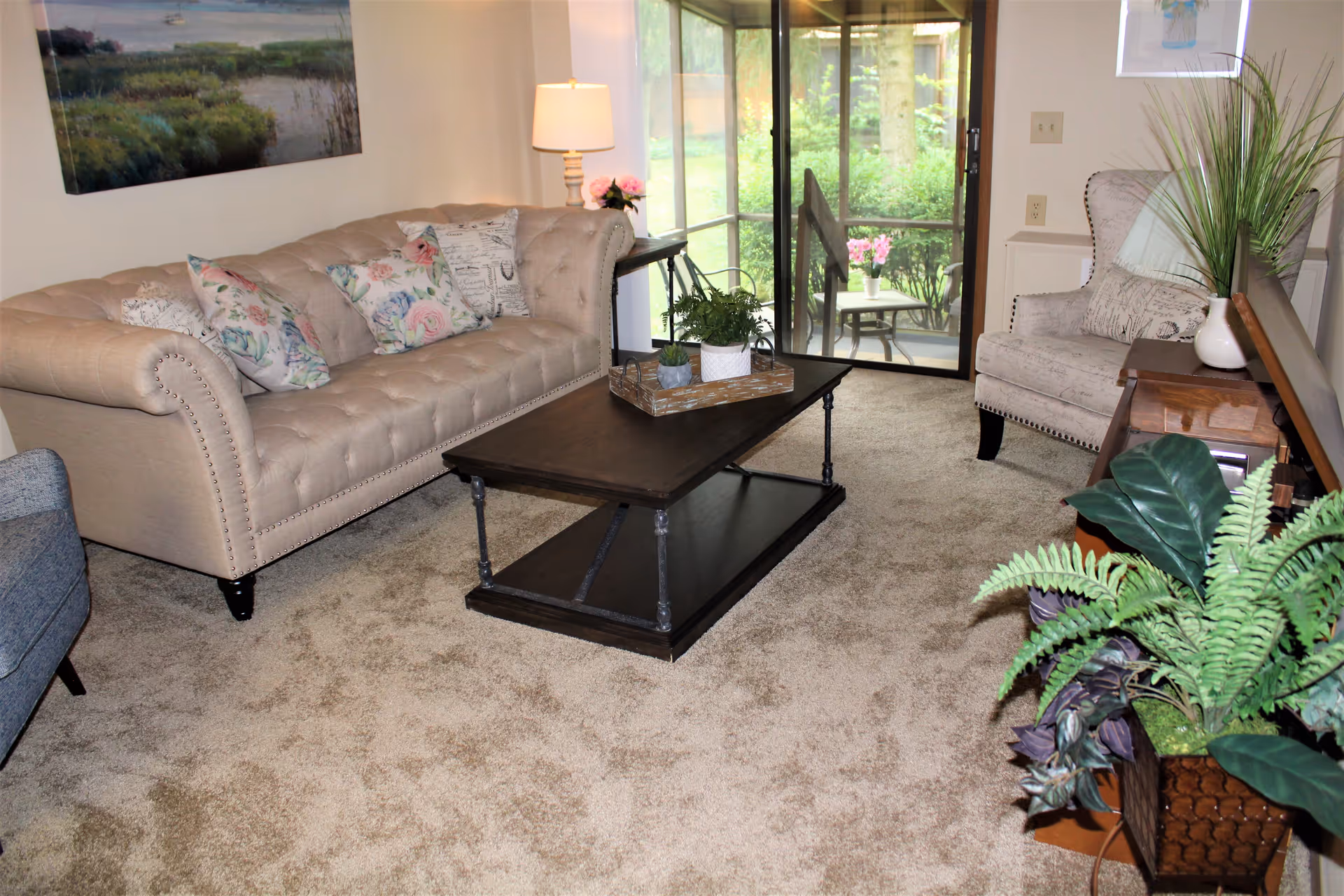 A cozy living room with a beige tufted sofa adorned with floral and patterned pillows, a dark wooden coffee table with a tray holding small plants, a beige armchair with a pillow, a side table with a lamp and flowers, and a sliding glass door leading to a patio with outdoor furniture. There are also several green plants in the foreground and a nature-themed painting on the wall.