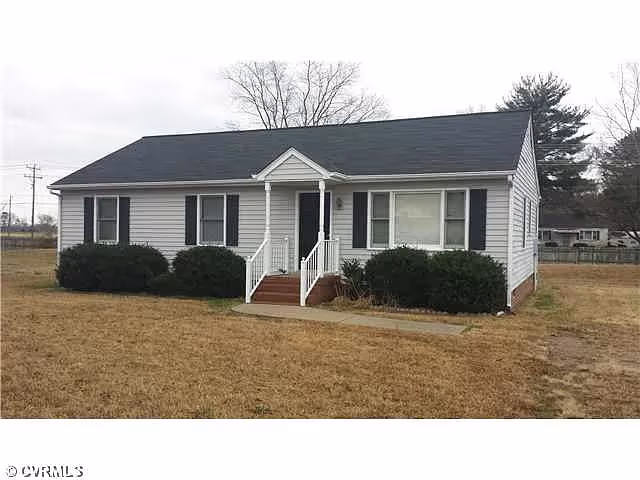 Single-story house with white siding, black shutters, and a small front porch with steps leading up to the entrance. The house is surrounded by a dry grass lawn and some bushes near the front.