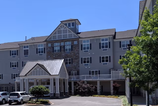 Exterior view of a multi-story senior living facility building with a covered entrance, several windows, and a small parking area with two cars. There are trees and shrubs around the building under a clear blue sky.