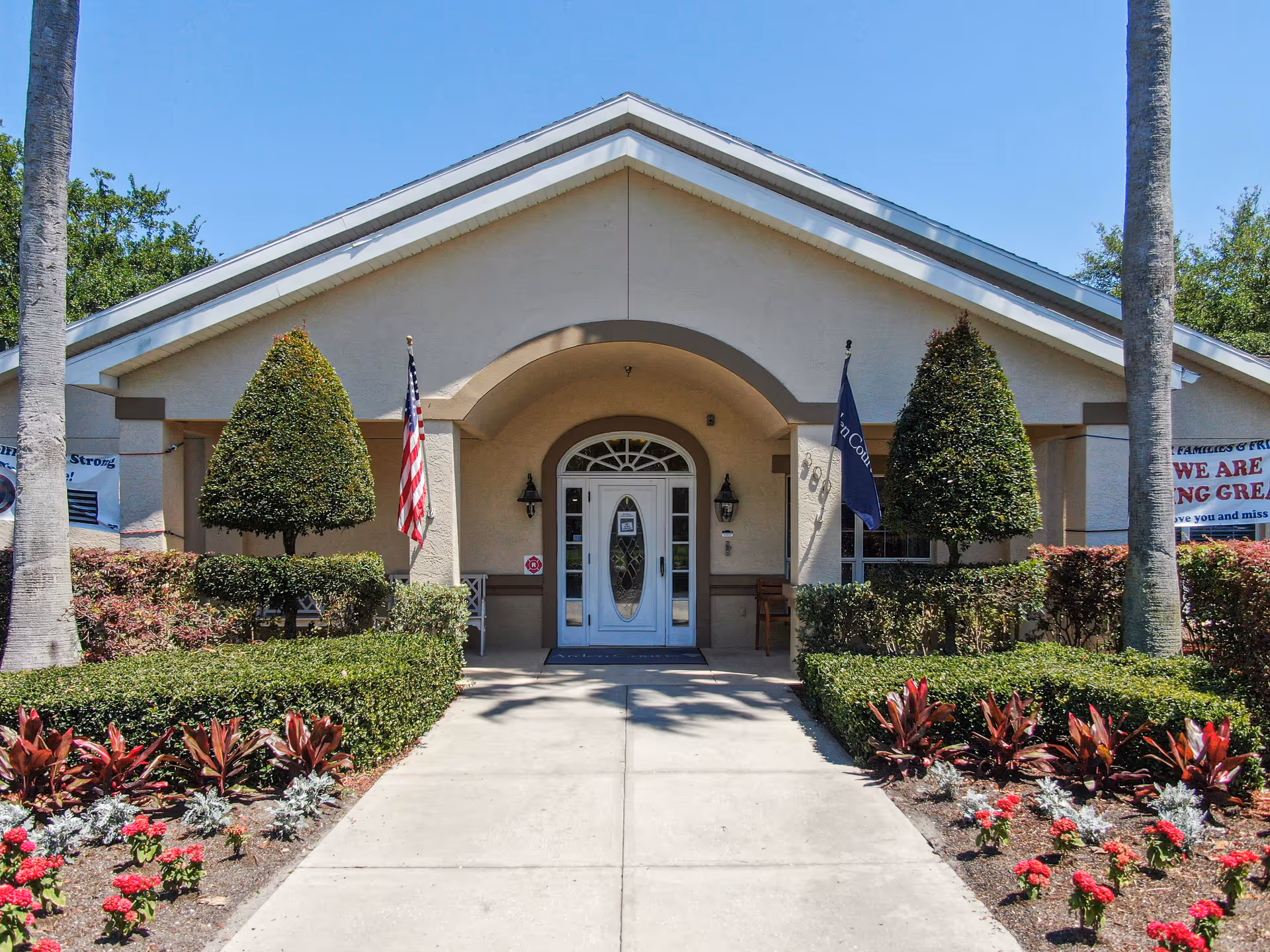 Front entrance of Arden Courts - ProMedica Memory Care Community in Largo, showing a beige building with a white door under an arched entryway, flanked by two neatly trimmed trees and American and facility flags. There are manicured bushes and flower beds along the walkway leading to the door.