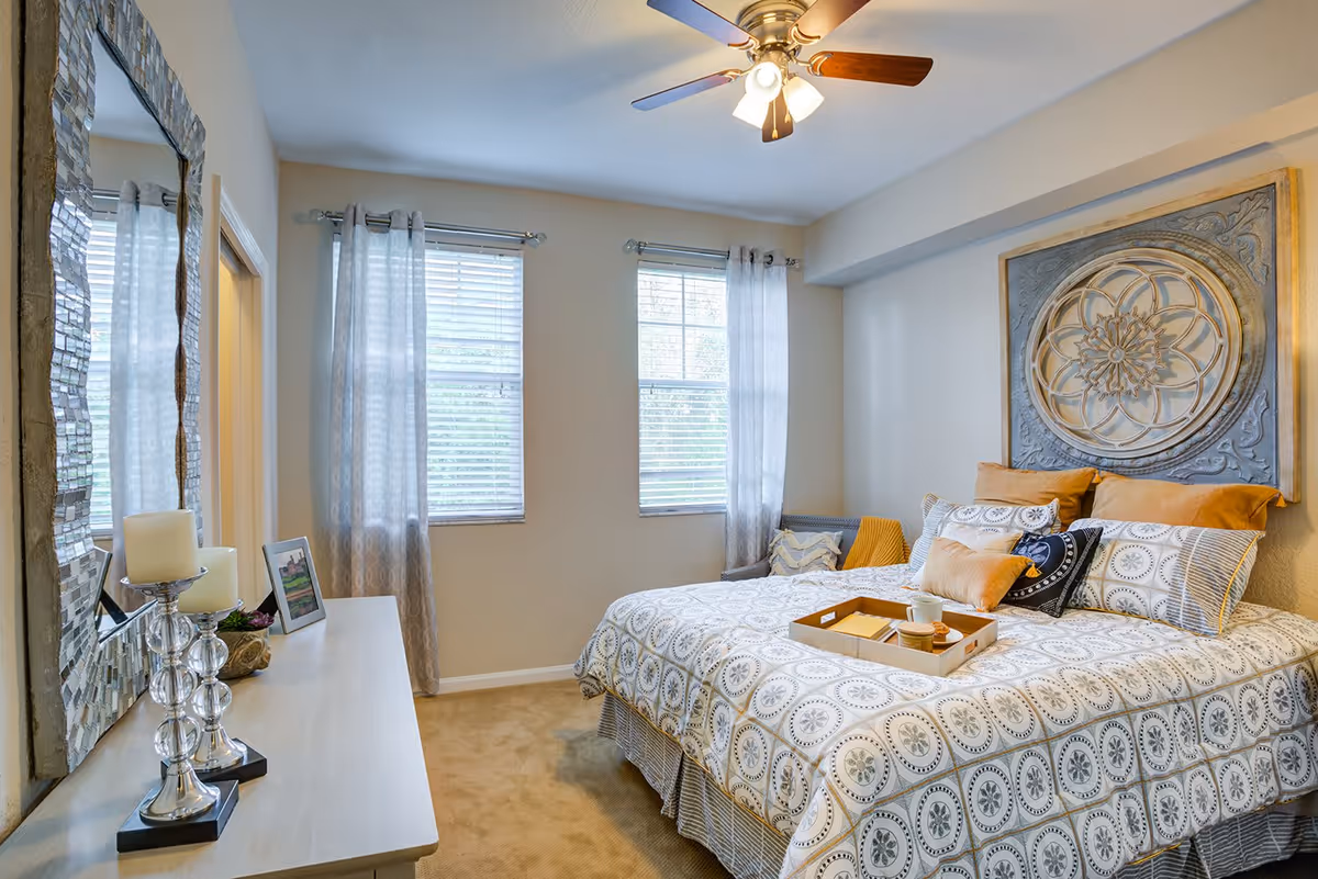 A cozy bedroom with a large bed covered in patterned bedding and multiple pillows. Above the bed is a decorative wall art piece. Two windows with sheer curtains allow natural light to fill the room. A ceiling fan with lights is mounted on the ceiling. A dresser with a large decorative mirror, candles, and a framed photo is positioned against one wall.