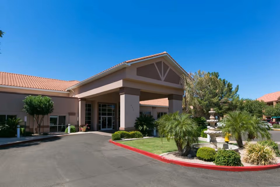 Exterior view of The Citadel Senior Living Community building entrance with a covered drop-off area, landscaped greenery including palm trees and bushes, a decorative fountain, and a clear blue sky.