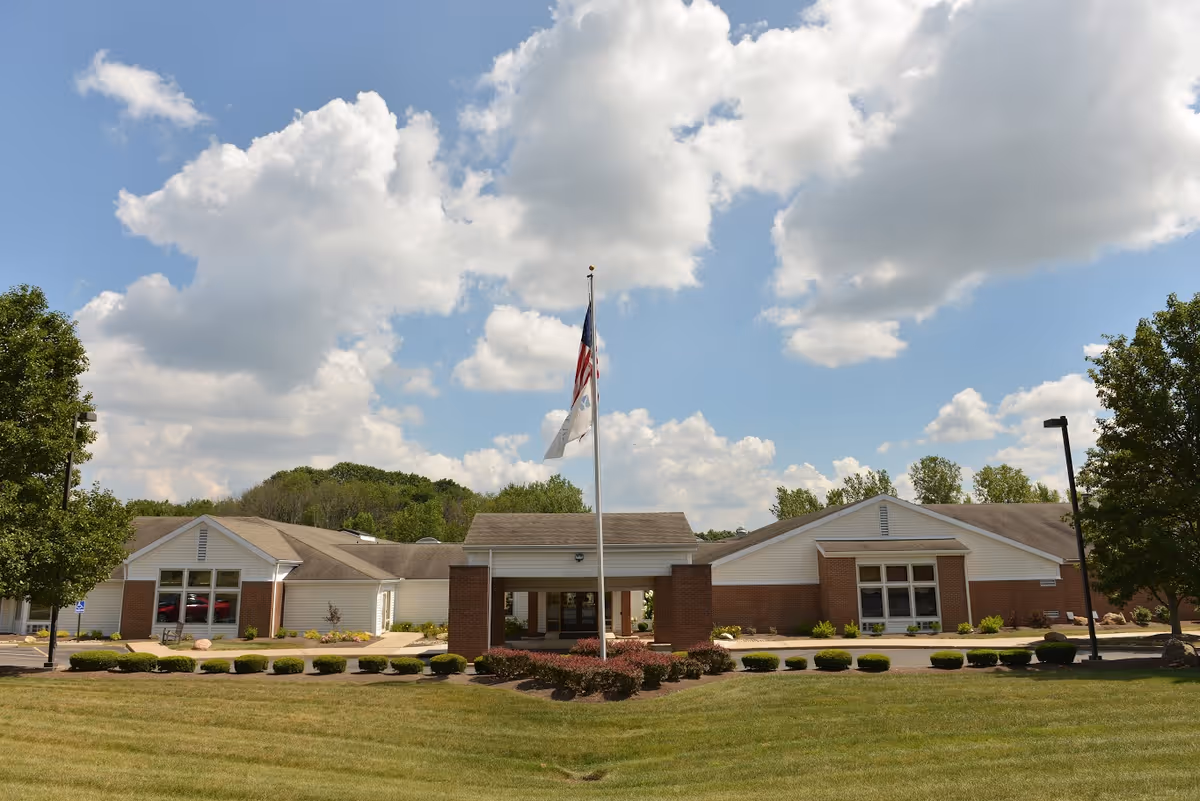 Exterior view of The Laurels of Massillon facility with a large lawn, neatly trimmed bushes, two trees, and a flagpole with the American flag and another flag in front of the building under a partly cloudy sky.