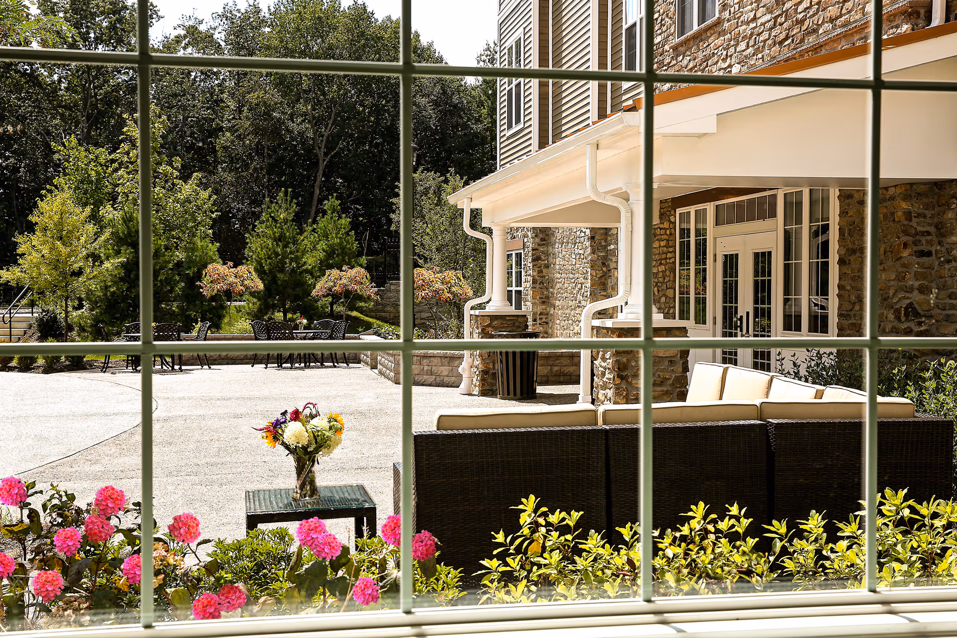 View through a window showing an outdoor patio area with cushioned wicker seating, a small table with a vase of flowers, pink flowers and green shrubs in the foreground, and a stone building with large windows and a covered porch. Trees and additional outdoor seating are visible in the background.