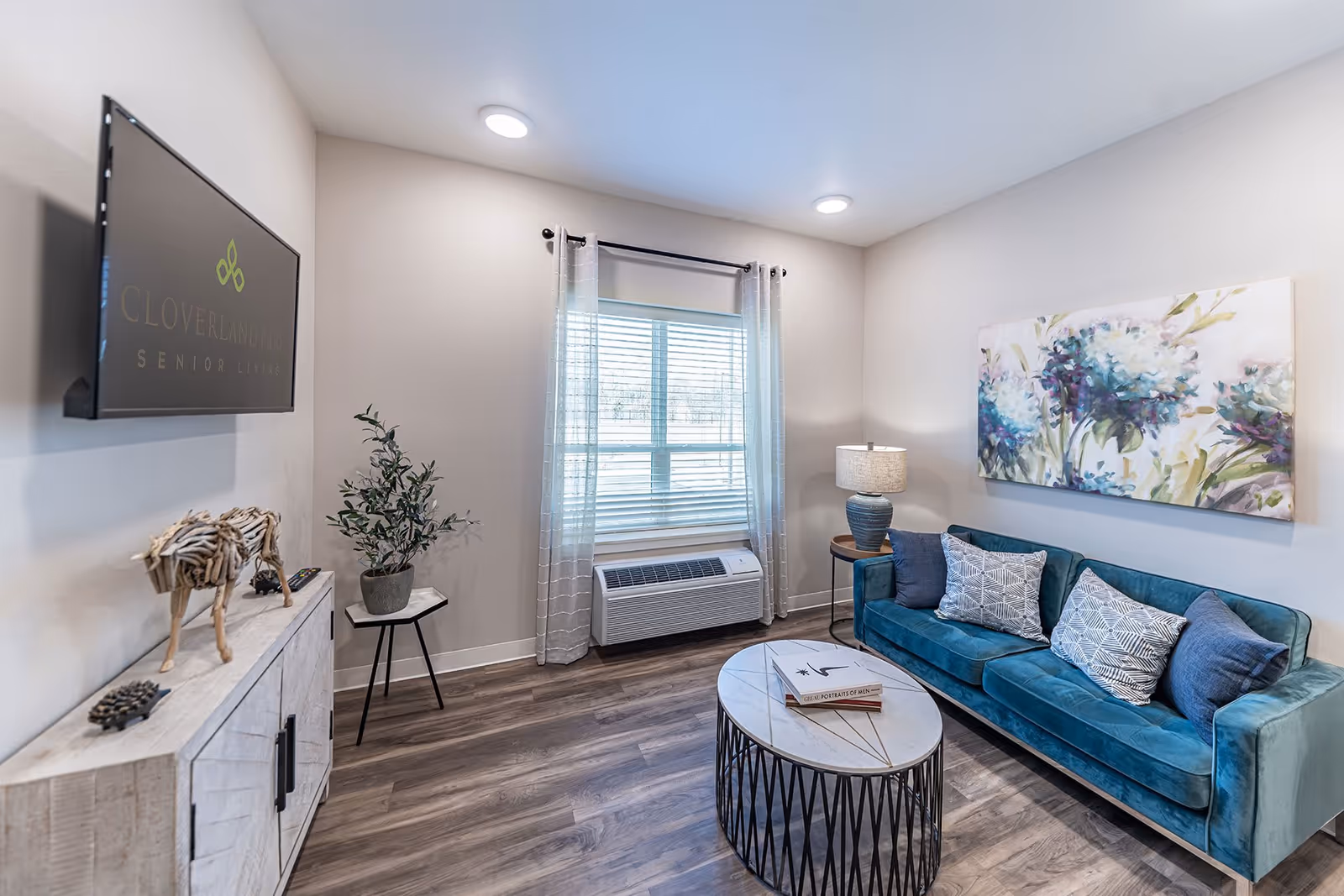 A cozy living room in Cloverland Park Senior Living featuring a teal velvet sofa with patterned cushions, a round marble-top coffee table with books, a side table with a lamp, a large floral painting on the wall, a window with sheer curtains, and a wooden cabinet with decorative items including a wooden animal sculpture and a potted plant.