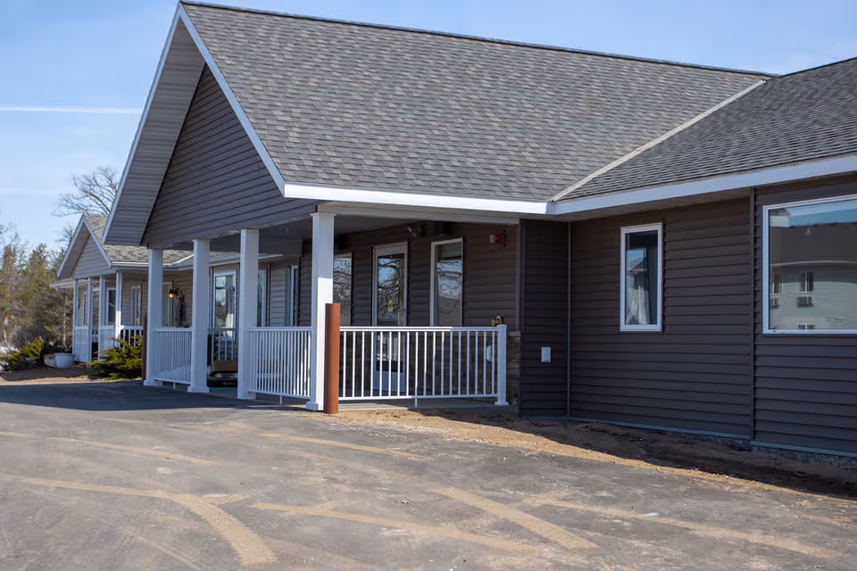 Exterior view of a single-story building with gray siding and a gabled roof. The building has a covered porch with white railings and columns, and several windows along the side. The ground in front is paved with visible tire marks.