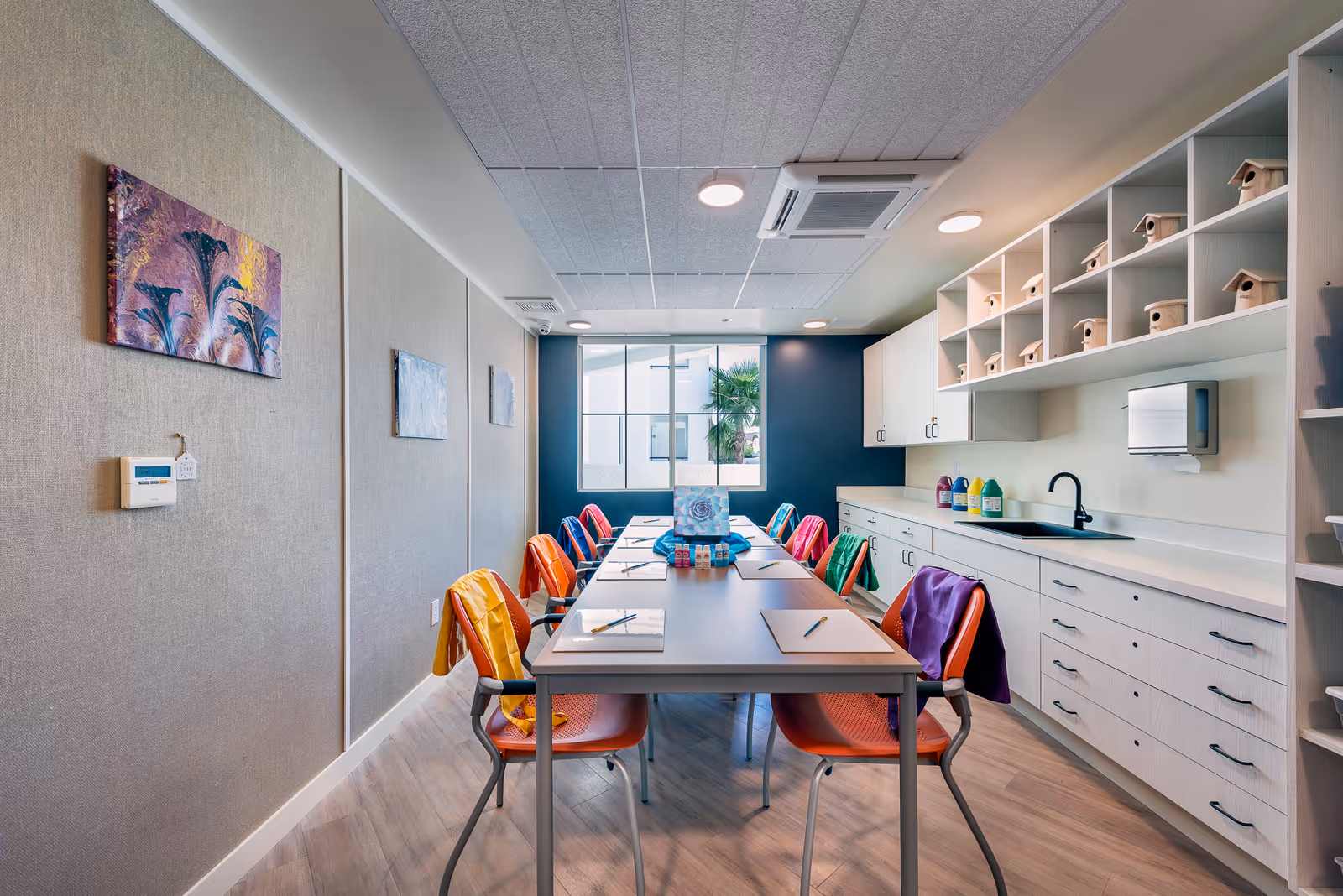 A bright and colorful activity room with a long table surrounded by orange chairs, each with a colorful jacket draped over the back. The table has papers, pencils, and art supplies arranged neatly. The room features light wood cabinetry with small birdhouses on the shelves, a sink, and a large window letting in natural light. The walls are decorated with paintings and the floor has a light wood finish.