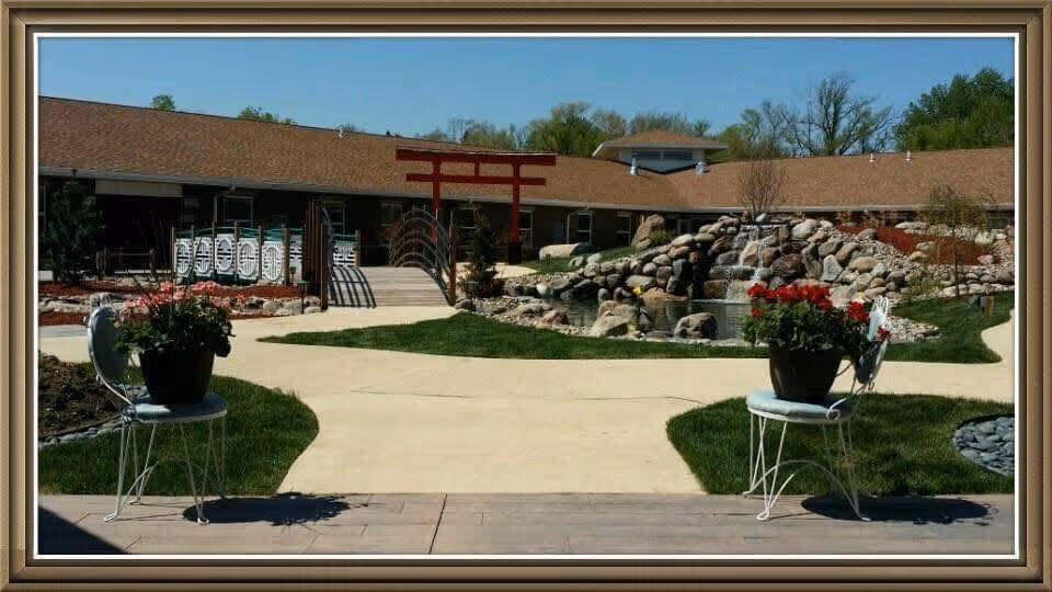 Outdoor garden area at Addington Place of Des Moines featuring a paved walkway, two white metal chairs with flower pots, a small bridge with decorative railings, a red torii gate, and a rock waterfall feature surrounded by greenery and a building in the background.