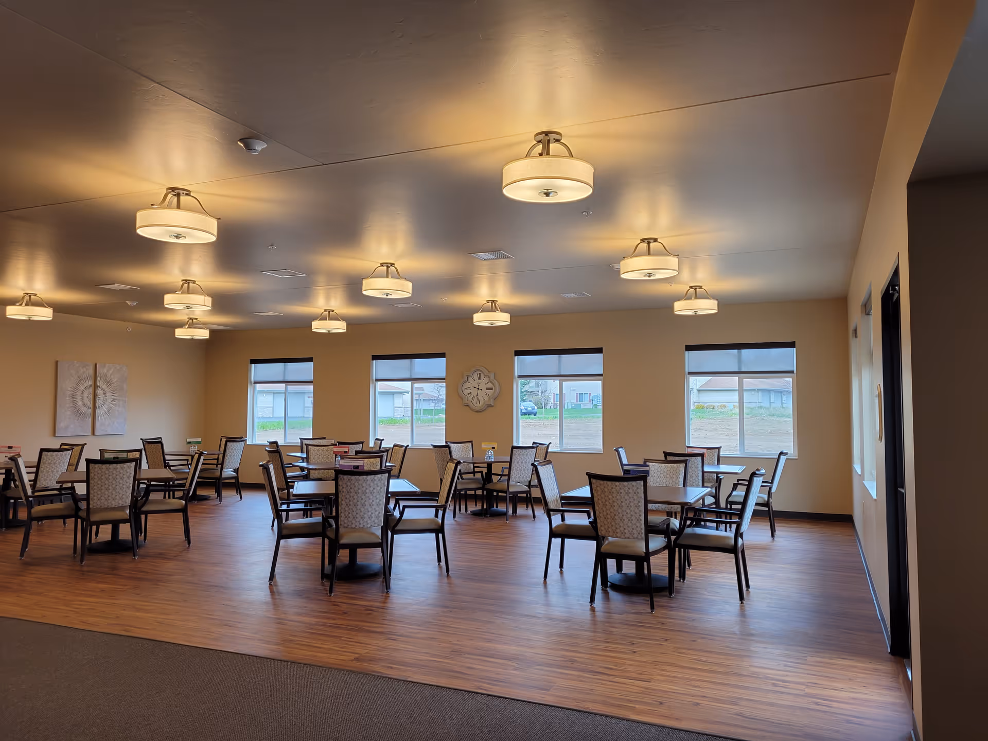 A spacious dining room with multiple square tables and cushioned chairs arranged neatly. The room has wooden flooring, beige walls, several windows letting in natural light, and ceiling-mounted light fixtures.