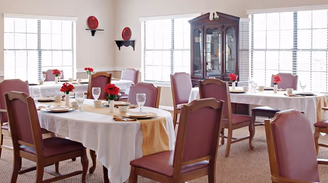 A bright dining room with several tables covered in white tablecloths and beige runners, each set with plates, glasses, and napkins. Red flowers in small vases decorate the tables. The room has large windows with white blinds, a wooden china cabinet, and wall shelves holding decorative red plates.