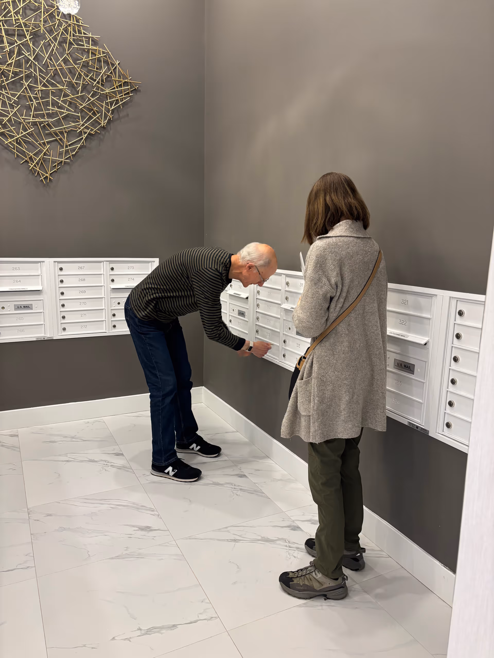 An elderly man and a woman standing in front of a wall of white mailboxes in a modern indoor mailroom with gray walls and white marble tile flooring. The man is bending down to open a mailbox while the woman stands nearby holding papers.