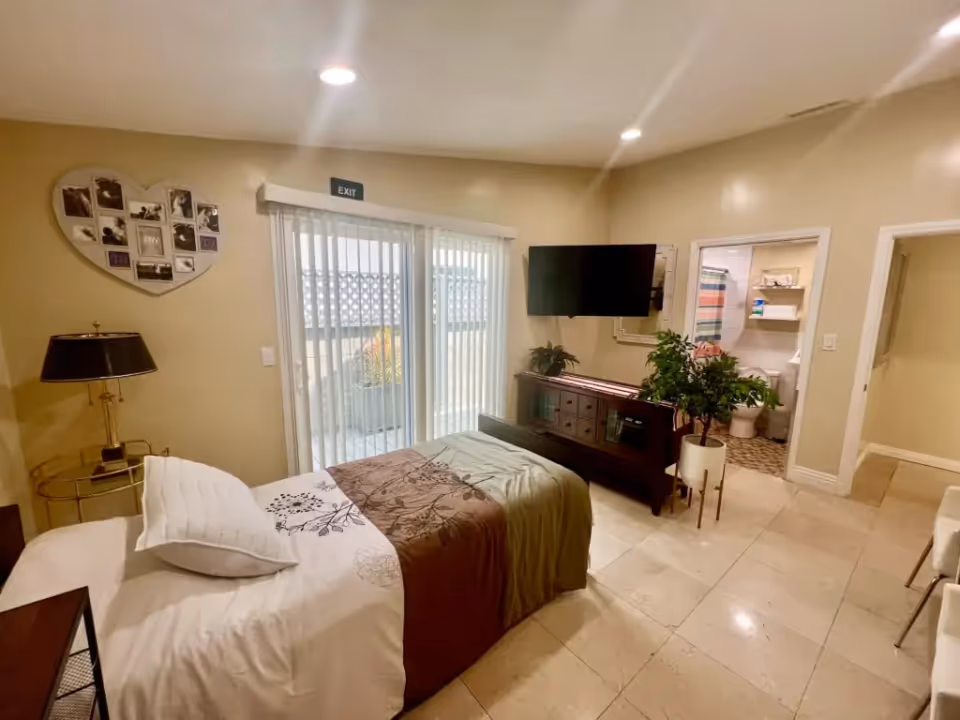 A cozy bedroom with a single bed covered in white and brown bedding, a bedside table with a lamp, a heart-shaped photo collage on the wall, a sliding glass door with vertical blinds leading outside, a flat-screen TV mounted on the wall above a wooden cabinet, a potted plant, and an open door revealing a bathroom with a toilet and shower curtain.