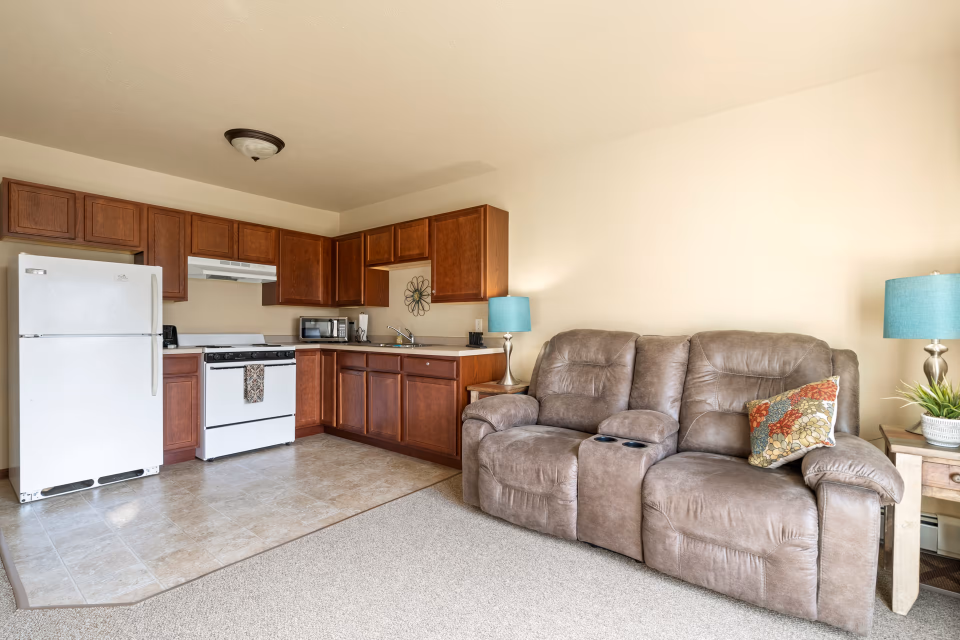 A small living area and kitchen in a senior living facility. The kitchen features wooden cabinets, a white refrigerator, stove, microwave, and a sink. Adjacent to the kitchen is a brown reclining loveseat with a colorful pillow and two blue lamps on side tables.