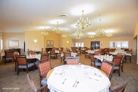 A spacious dining room with multiple round and rectangular tables set with white tablecloths, glasses, and silverware. The room features several chandeliers hanging from the ceiling, beige walls, and framed artwork. Chairs with patterned upholstery surround each table.