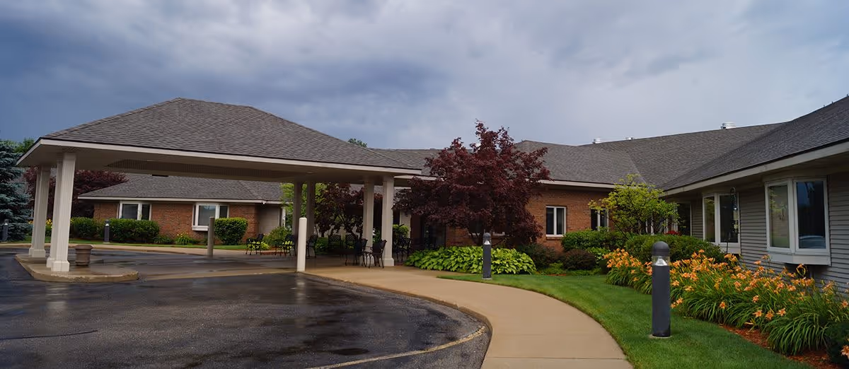 Exterior view of Sheldon Meadows Assisted Living Center showing a covered entrance with a driveway and a sidewalk leading to the building. The building is surrounded by well-maintained landscaping including bushes, trees, and flowers under a cloudy sky.
