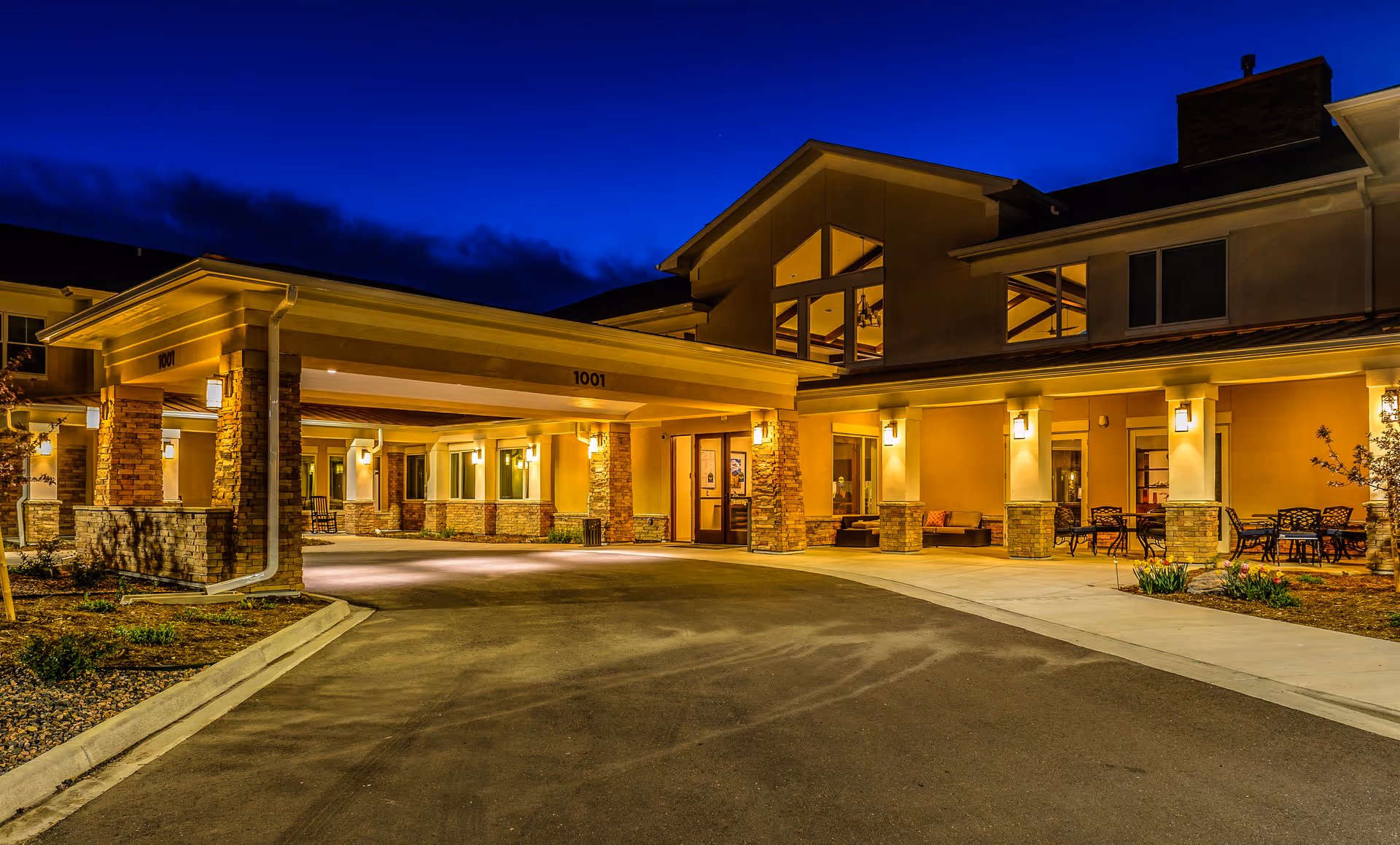 Exterior nighttime view of AltaVita Assisted Living facility entrance with warm lighting, covered drop-off area, and outdoor seating on the right side.