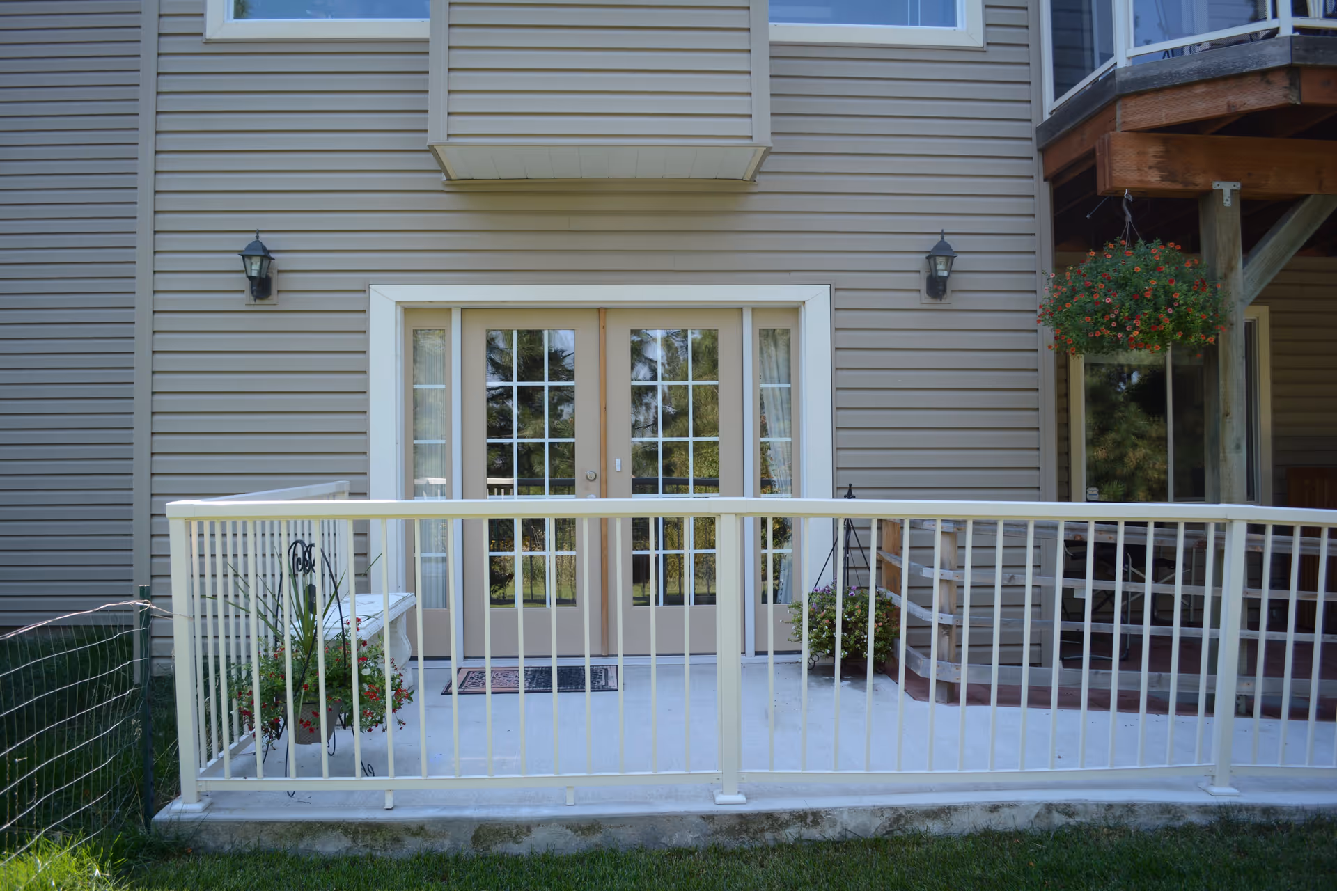 Exterior view of a beige building with a small concrete patio enclosed by a white metal railing. The patio has two potted plants with flowers, a white bench, and double glass doors with white frames leading inside. There are two wall-mounted lantern-style lights on either side of the doors and a hanging flower basket on the right side under a wooden deck.
