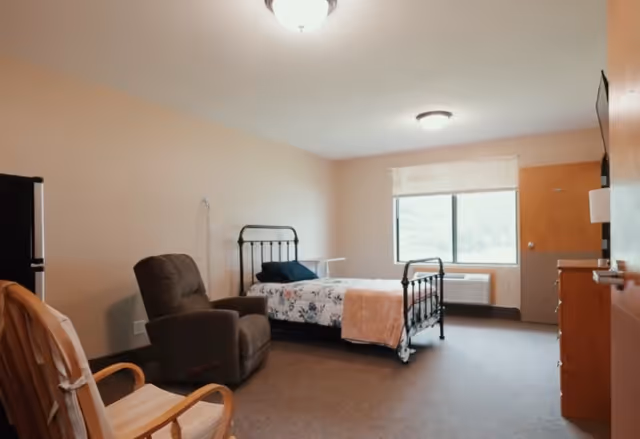 A simple and clean assisted living bedroom with a single metal bed frame, floral bedding, a brown recliner chair, a wooden rocking chair, a window with a white blind, a small refrigerator, and a dresser with a TV mounted above it.