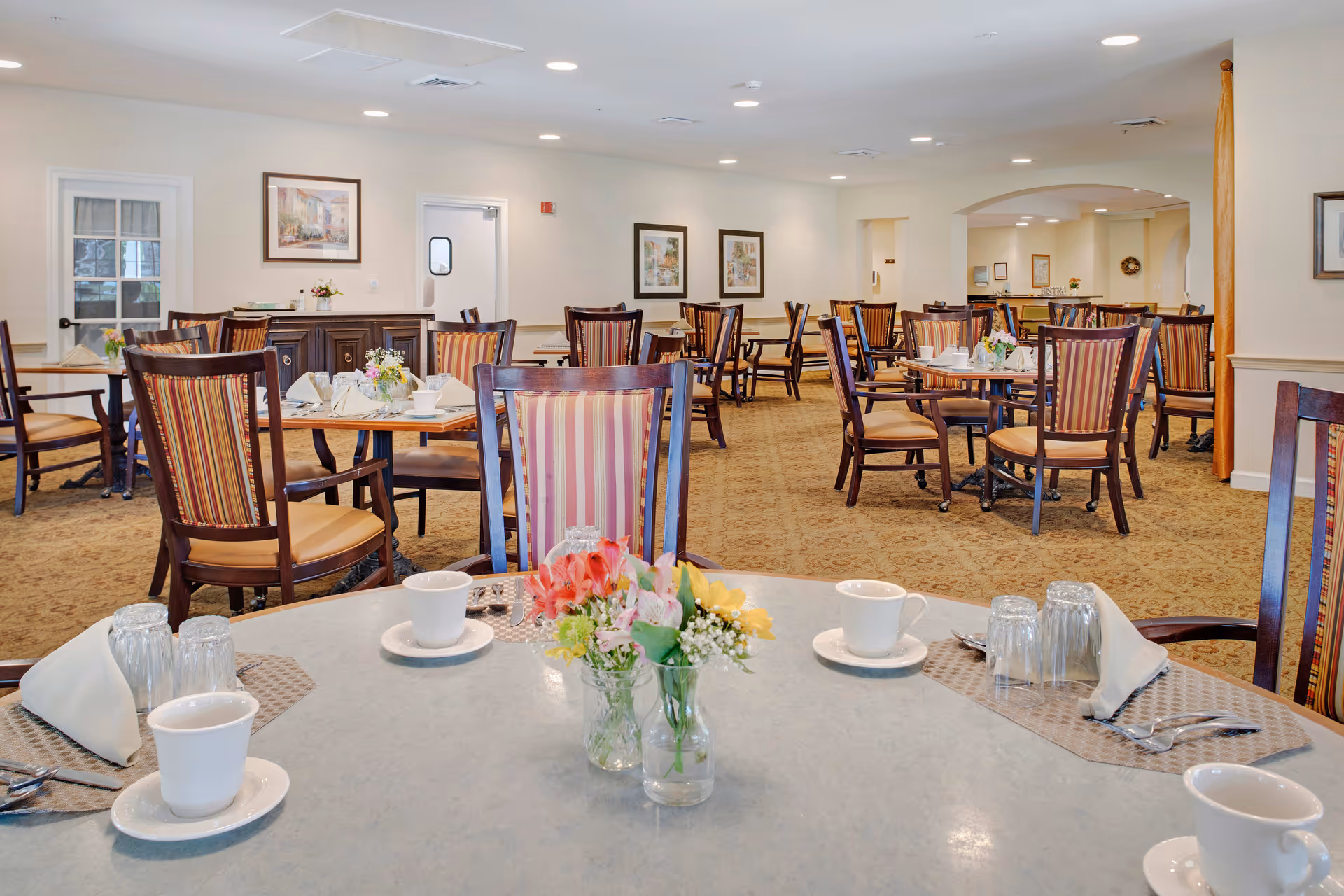 A spacious dining room with multiple wooden tables and chairs featuring striped upholstery. Tables are set with white cups, saucers, glasses, silverware, and napkins. Small floral centerpieces decorate the tables. The room has beige walls adorned with framed artwork and a carpeted floor. Bright ceiling lights illuminate the area.