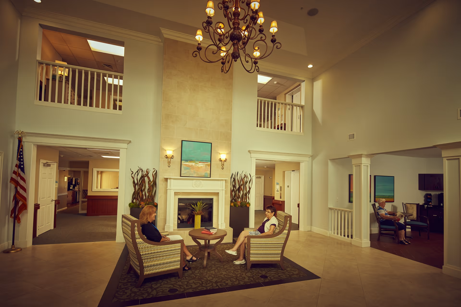 A spacious, well-lit senior living facility common area with high ceilings and a chandelier. Two women are seated in cushioned armchairs around a small round table with a plant and books. There is a fireplace with a painting above it, decorative plants on either side, and an American flag to the left. Another elderly woman is seated in a separate seating area to the right, reading a newspaper.